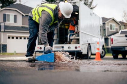 Utility worker in a reflective vest repairs a blue water hose as water gushes onto a residential street.