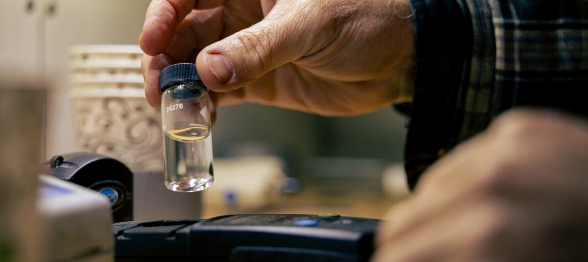 A person's hand holds a small glass bottle of water that will be tested.