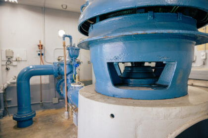 Close-up of a large worn blue industrial valve or pump housing with exposed dark internal machinery, surrounded by pipes in a mechanical room.