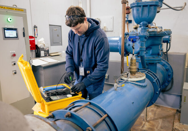 Worker in safety glasses tests industrial equipment beside large blue pipes using instruments in an open yellow case.