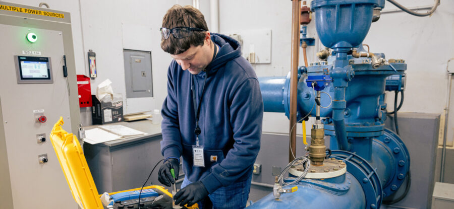 Worker in safety glasses tests industrial equipment beside large blue pipes using instruments in an open yellow case.