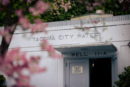 White Tacoma City Water facility labeled “WELL 11-A,” partly obscured by blurred pink blossoms.