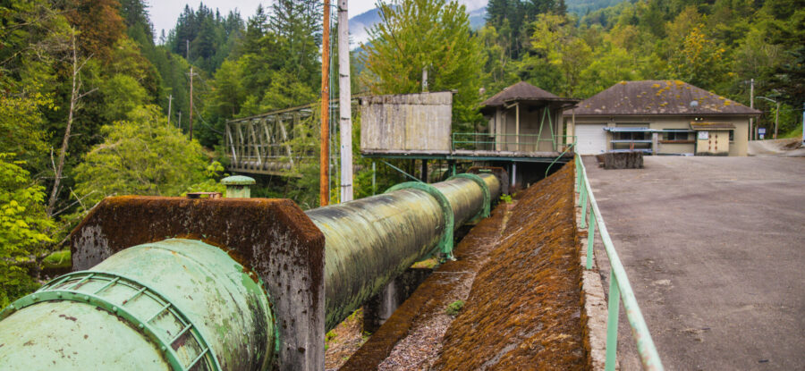 Large moss-covered green water pipe beside a utility building in a forested, misty mountain watershed.