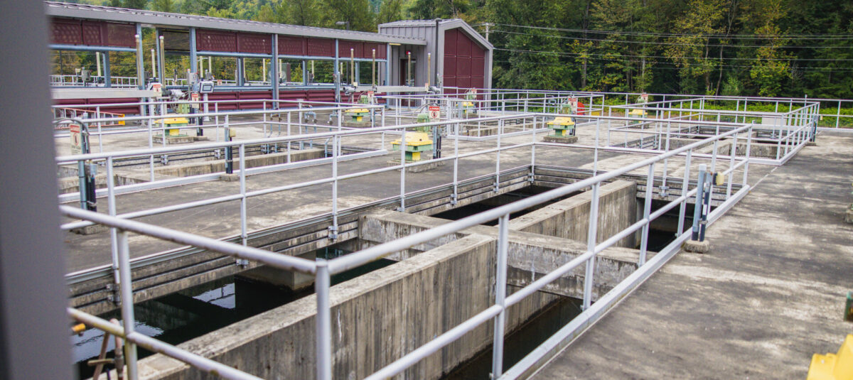 Water treatment facility with concrete water channels, pipes, and safety railings against a forested, misty mountain backdrop.