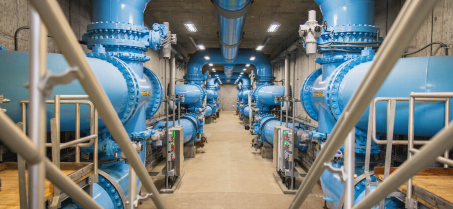 Interior of a water treatment facility with large blue pipes, valves, and filtration equipment arranged along a central walkway.