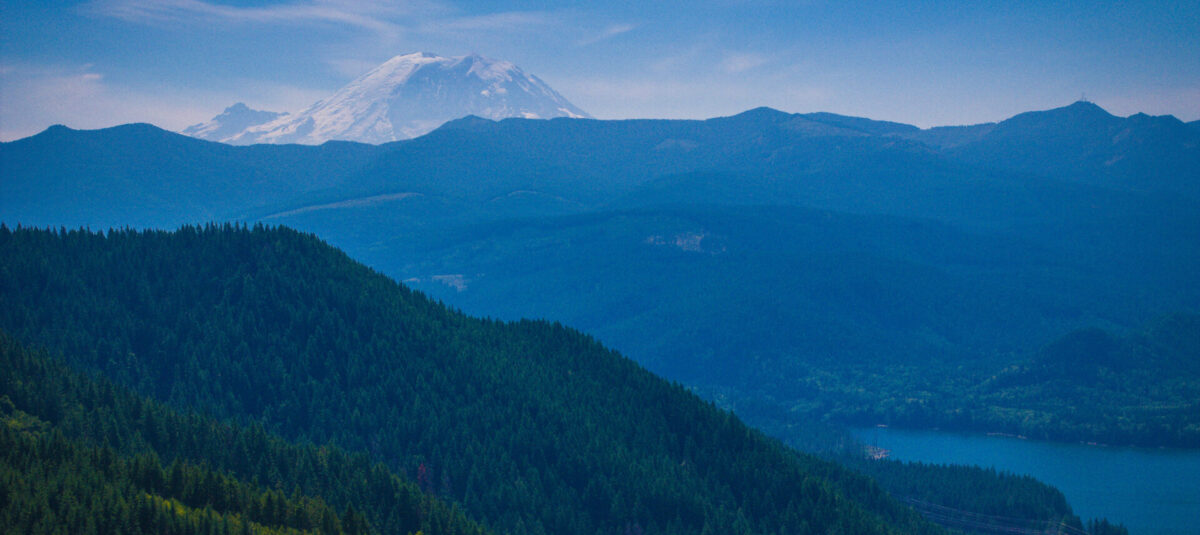 Snow-covered Mount Rainier rises behind forested hills and the Green River watershed, with a blue lake visible at lower right.