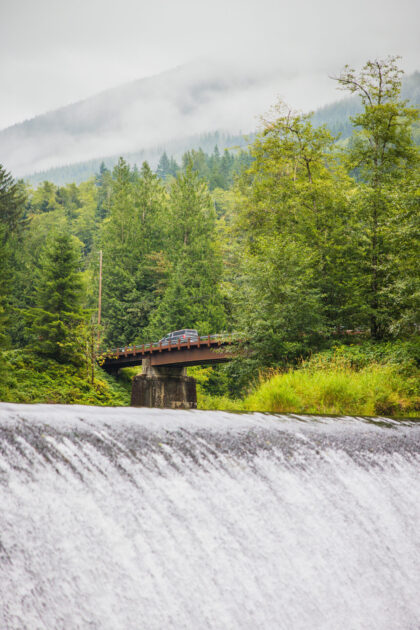A misty, forested mountain landscape with a vehicle crossing a bridge over a rushing river.