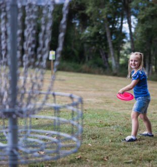 young girl in blue shirt throwing a frisbee in disc golf