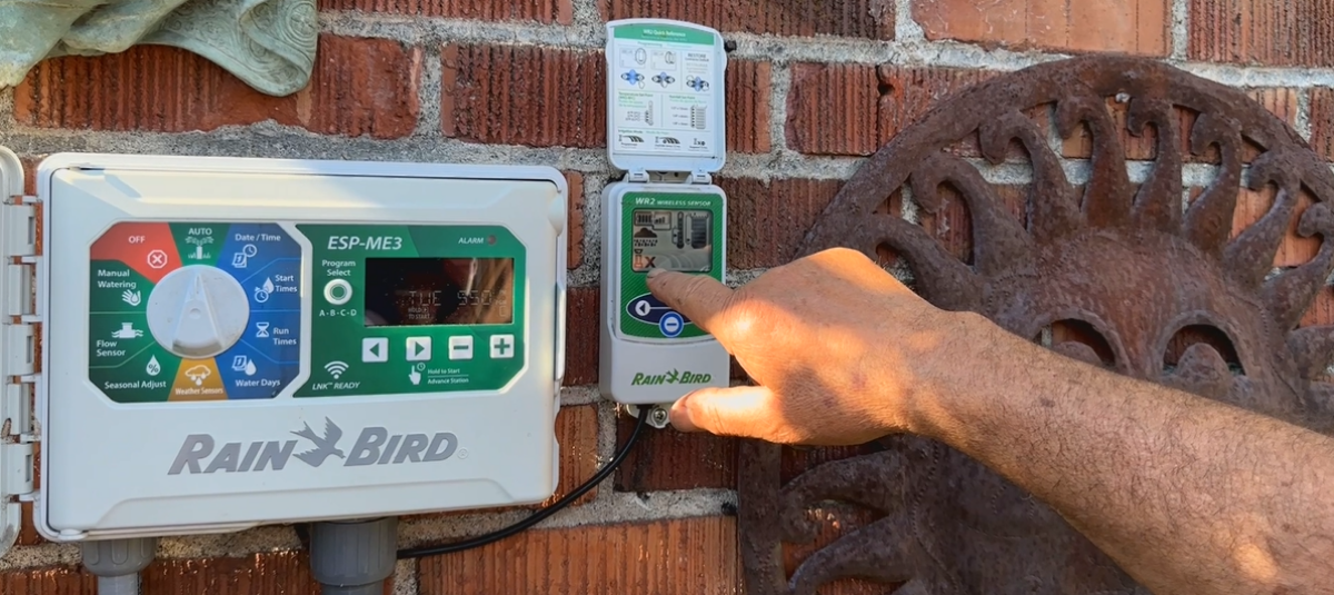A hand points to a Rain Bird WR2 wireless sensor mounted beside an ESP-ME3 irrigation controller on a brick wall.