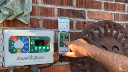 A hand points to a Rain Bird WR2 wireless sensor mounted beside an ESP-ME3 irrigation controller on a brick wall.